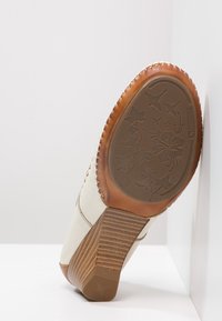 Valkoiset nahkakengät, joissa on ruskea ommeltu pohja, ja joissa on teksturoitu kanta sekä kukkakuvio pohjassa. Huomattavat kontrastivärit ja materiaalit.