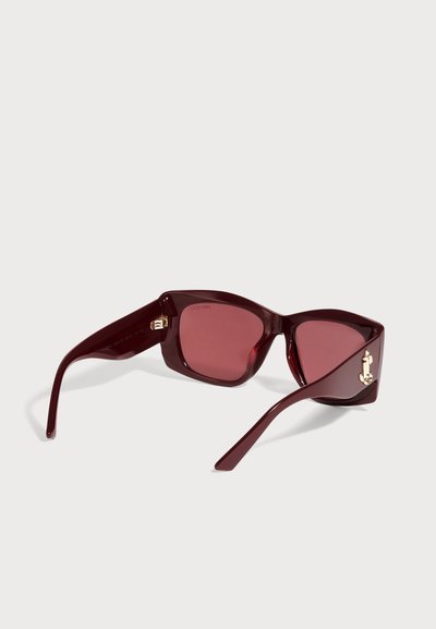 Lunettes de soleil bordeaux en forme œil-de-chat, avec des verres teintés rose et des détails dorés, présentées de biais sur un fond clair uni.
