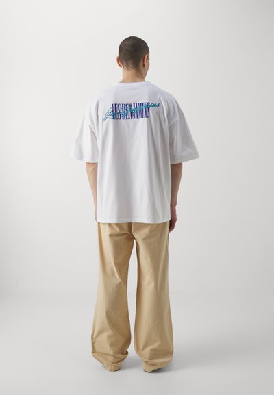 T-shirt blanc oversized avec du texte coloré dans le dos, associé à un pantalon beige ample, mettant en valeur une coupe décontractée et un style casual.