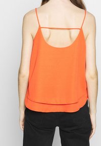 Orange ärmlös topp med rundad halsringning, som har tunna spagettiband och ett lagerat nederkant. Slät textur, avslappnad design.