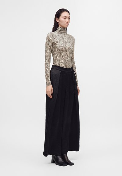 Femme portant un col roulé beige à manches longues imprimé serpent, un pantalon large noir taille haute et des bottines noires, debout sur un fond blanc.