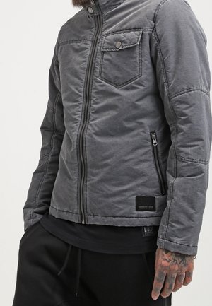 Homme portant une veste matelassée grise zippée avec poche poitrine et fermeture éclair noire, associée à un pantalon noir, montrant une main tatouée.