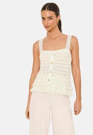 Vrouw draagt een crèmekleurige gehaakte mouwloze top met knopen en een gerimpelde zoom, gecombineerd met lichtbeige wijde broek.