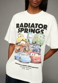 Bílý tričko s grafikou karikaturních aut a textem "Radiator Springs". Oversized střih, měkká tkanina a barevný design.
