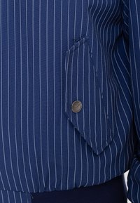 Primo piano di tessuto blu a righe sottili con tasca a patta fissata da un bottone a pressione metallico rotondo.