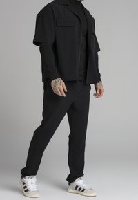 Svart kortärmad zip-up skjorta, slim-fit svarta byxor, silver- och svarta sneakers med tre ränder som visar en slät textur och minimalistisk design.