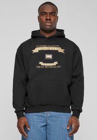 Black hoodie featuring a gold graphic with "KARL KANI" and a trademark label. It has a front pocket and ribbed cuffs and hem.