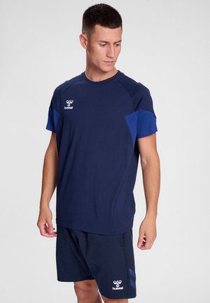 Mężczyzna w granatowej sportowej koszulce i szortach Hummel, patrzący w dół i na bok na jednolitym jasnym tle.