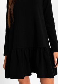 Czarna sukienka z długimi rękawami, uszyta z miękkiego materiału, z rozkloszowanym dołem ozdobionym falbanką, luźny krój i prosty, minimalistyczny design.