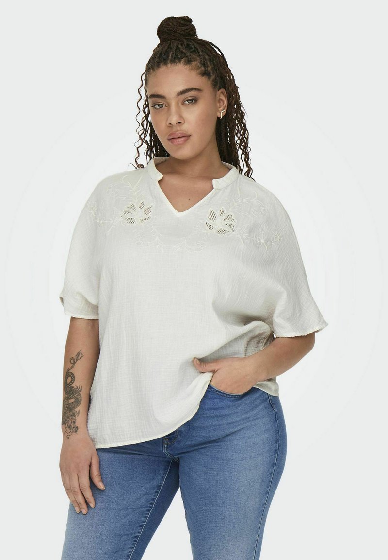 Hvid kortærmet top med en tekstureret overflade, der har broderede blomsterdetaljer ved halsudskæringen, parret med blå jeans.