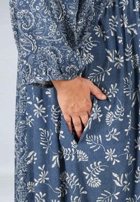 Denim jurk met een donkerblauwe basis en een witte bloemen paisley patroon. Heeft lange mouwen en een zijzak die wordt benadrukt door een hand.