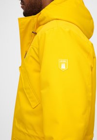 Geel waterdichte jas met capuchon, met een voorzak en een wit logo-opdruk op de mouw. Gladde textuur, modern ontwerp.