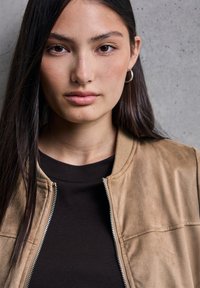 Giubbotto bomber in suede marrone con zip frontale e colletto a coste, indossato sopra una maglietta nera. Capelli lunghi e lunghi, scuri, che incorniciano il viso.