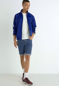 Giacca blu con zip, t-shirt bianca, pantaloncini di jeans arrotolati e sneakers bordeaux con dettagli bianchi. La modella è in piedi di fronte a uno sfondo neutro.