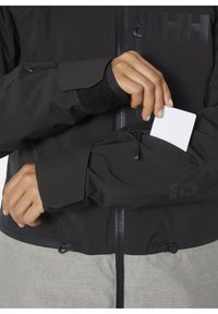Schwarze wasserdichte Jacke mit einer Reißverschlusstasche am Arm, Klettverschlussmanschette und grauem unteren Abschnitt. Mit Logo und verstellbarem Saum mit Zugbändern.