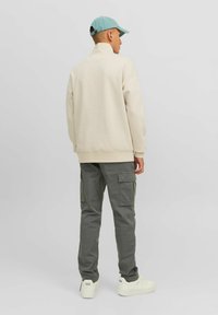 Beige Sweatshirt mit hohem Kragen und graue Cargo-Hose mit Taschen. Das Modell trägt eine hellblaue Mütze und weiße Sneaker.