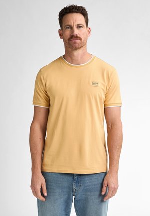 Gelbes Baumwoll-T-Shirt mit kurzen Ärmeln, weißem Kragen und Ärmelabschlüssen, mit einem kleinen quadratischen Logo-Patch auf der Brust. Tragen mit blauen Jeans.