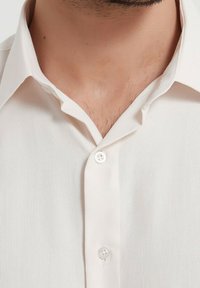 Chemise courte à manches courtes beige clair avec un tissu texturé, deux boutons à l'avant et un col classique, mettant l'accent sur la simplicité et la fonctionnalité.