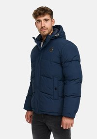 Mężczyzna w granatowej kurtce puchowej z kapturem, stojący na tle jednolitego białego tła, zwrócony lekko w lewo.