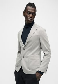 Άνδρας που φοράει ανοιχτό γκρι σακάκι πάνω από μαύρο ζιβάγκο, στέκεται με το ένα χέρι στην τσέπη, κοιτάζοντας προς τα κάτω δεξιά του, μπροστά σε απλό φόντο.