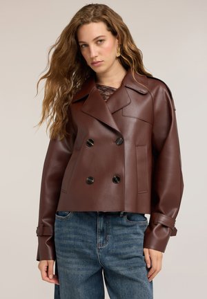 Femme aux longs cheveux ondulés portant une veste en cuir marron foncé à double boutonnage et un jean bleu sur un fond beige.