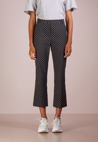 Person som bär svarta korta byxor med små blommönster, grå t-shirt och vita sneakers, står på en slät golv mot en enfärgad bakgrund.