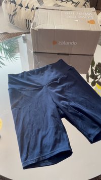 Sötétkék sportnadrág sima, rugalmas anyagból; széles derékrésszel és combközép hosszúsággal rendelkezik. A háttérben egy kartondoboz található.