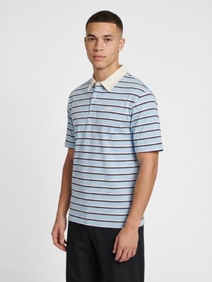 Junger Mann trägt ein hellblau und schwarz gestreiftes Poloshirt mit weißem Kragen und schwarzen Hosen und steht vor einem schlichten weißen Hintergrund.