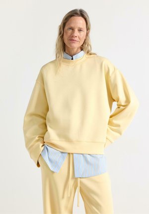 Kvinde med lyst hår iført en lysegul sweatshirt og bukser over en blå- og hvidstribet skjorte med knapper, stående med hænderne i lommerne.