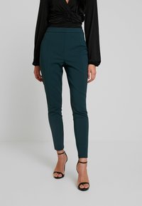 Teal, figurnära byxor med en mjuk textur och en söm fram, tillsammans med svarta, remmade klackar och en långärmad svart topp.