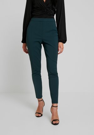 Teal, slim-fit broek met een gladde textuur en een detail van een voornaad, gecombineerd met zwarte sandaaltjes met een bandje en een zwart longsleeve top.