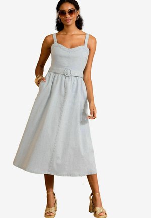 Hellblaues, gestreiftes Midi-Kleid mit tailliertem Oberteil, breiten Trägern, einem Gürtel mit einer runden Schnalle und seitlichen Taschen. Dazu werden Wedge-Sandalen getragen.