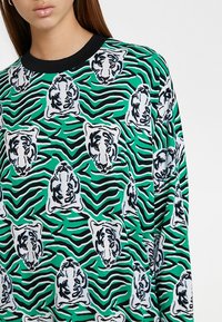 Långärmad topp med ett grönt och svart tigerrandigt mönster som har vita tigerhuvuden. Tillverkad av mjukt tyg med klassisk rundad hals.