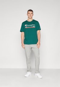 Camiseta de algodón verde con el logotipo "Champion New York" en blanco y rojo, combinada con pantalones de chándal grises claros y zapatillas blancas. Conjunto simple y casual.