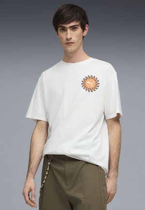 Młody mężczyzna o krótkich ciemnych włosach, ubrany w białą koszulkę z pomarańczowym nadrukiem słońca oraz oliwkowozielone spodnie z breloczkiem z koralików.