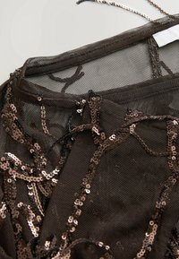 Gros plan sur un tissu noir transparent avec des paillettes brunes tourbillonnantes et un collier chaîne en métal fin sur une surface claire.