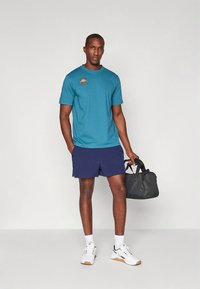 Žydros sportinės marškinėliai su oranžiniu logotipu, derinami su tamsiai mėlynais šortais. Aprangą papildo balti sportbačiai ir juoda sportinė kuprinė.