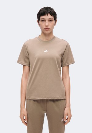 Osoba z krótkimi ciemnymi włosami, ubrana w beżowy pasujący t-shirt i spodnie z małym białym logo na piersi i udzie, stoi zwrócona przodem.