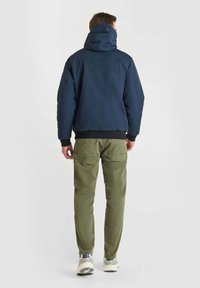 Giacca con cappuccio blu navy con design imbottito, polsini e orlo a costine, abbinata a pantaloni verde oliva e sneaker grigie.