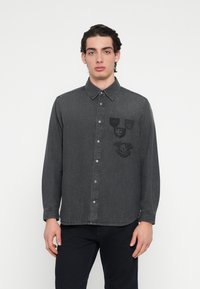 Πουκάμισο από γκρι τζιν με μακριά μανίκια και γιακά με κουμπιά, με τρία μαύρα κεντημένα σχέδια στο αριστερό μέρος του στήθους και ασημένια κουμπιά.