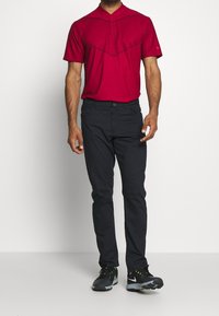 Röd pikétröja med krage och strukturerat mönster, kombinerad med mörka byxor och svarta träningsskor med gröna accenter.