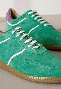 Une paire de baskets en suède vert avec des rayures blanches, des lacets à motifs blancs, des empiècements de talon violets et des semelles en caoutchouc marron sur une surface beige.