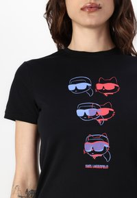 Černé bavlněné tričko s grafickým potiskem stylizovaných postav v brýlích v modré a červené barvě. Text "KARL LAGERFELD" na spodním okraji.