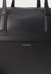 Sac à main en cuir noir avec une texture grainée, des poignées supérieures et un subtil logo Calvin Klein en argent à l'avant. Design simple et élégant.