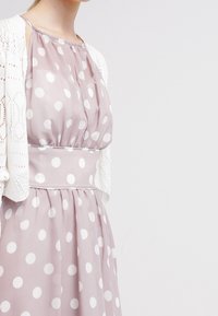 Světle růžové šaty s puntíky a zúženým pasem, doplněné průsvitným topem a bílým krajkovým kardiganem. Látka má jemnou, splývavou texturu.