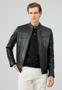 Zwarte leren jas met een staande kraag, rits aan de voorkant en twee borstzakken. Heeft een gladde textuur en een getailleerd ontwerp.