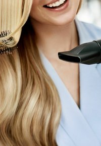 Asciugacapelli con un beccuccio nero, posizionato vicino a lunghi e lisci capelli biondi, che vengono pettinati con una spazzola rotonda a setole nere.