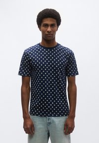 Μπλούζα μπλε ναυτικού χρώματος με λευκά κυκλικά σχέδια, στρογγυλή λαιμόκοψη, κοντά μανίκια, κατασκευασμένη από μαλακό υλικό, φορεμένη με ανοιχτόχρωμο τζιν παντελόνι.