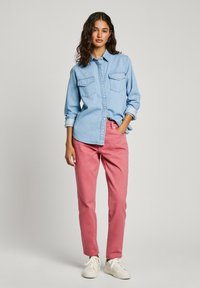 Lichtblauwe denim overhemd met twee borstzakken, gecombineerd met roze broek en witte sneakers. Het model staat tegen een effen achtergrond.