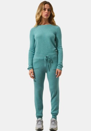 Completo loungewear in maglia turchese con maniche lunghe e vita con cordino. Texture morbida, design aderente, abbinato a sneakers grigie.
